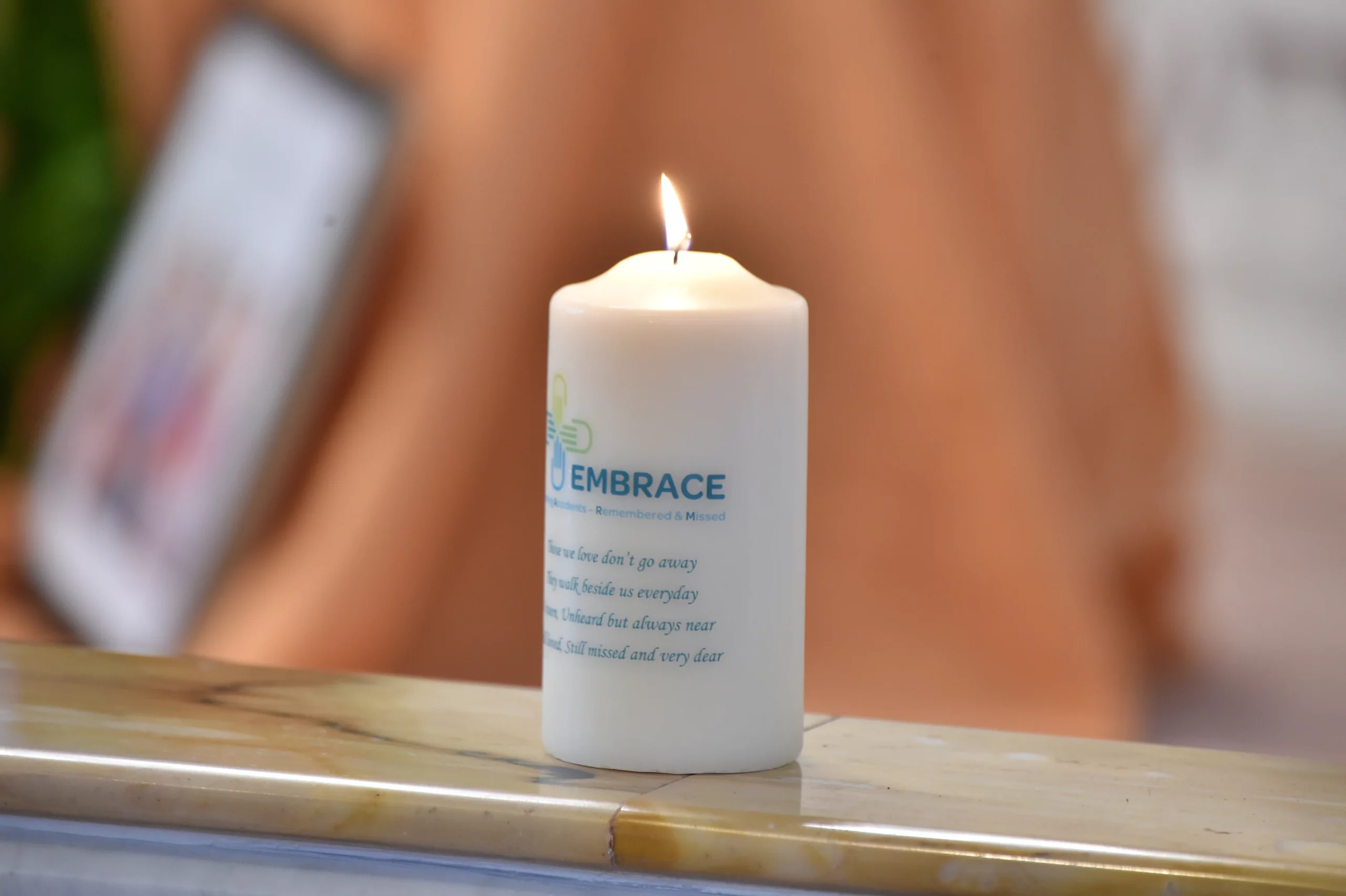 Remembrance Service White candle with 'EMBRACE' written on it, placed on a marble surface